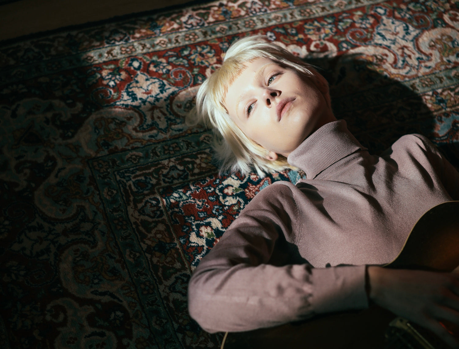 blonde girl laying down on a persain carpet playing guitar.